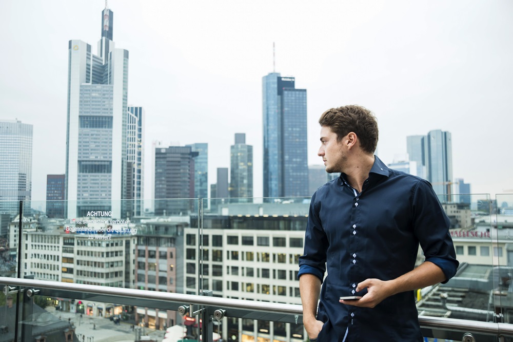 Mann vor der Skyline von Frankfurt mit Smartphone in der Hand blickt in die Ferne